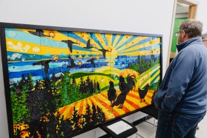 Une personne admire une mosaïque collective, qui illustre un paysage avec un soleil, la nature, des animaux